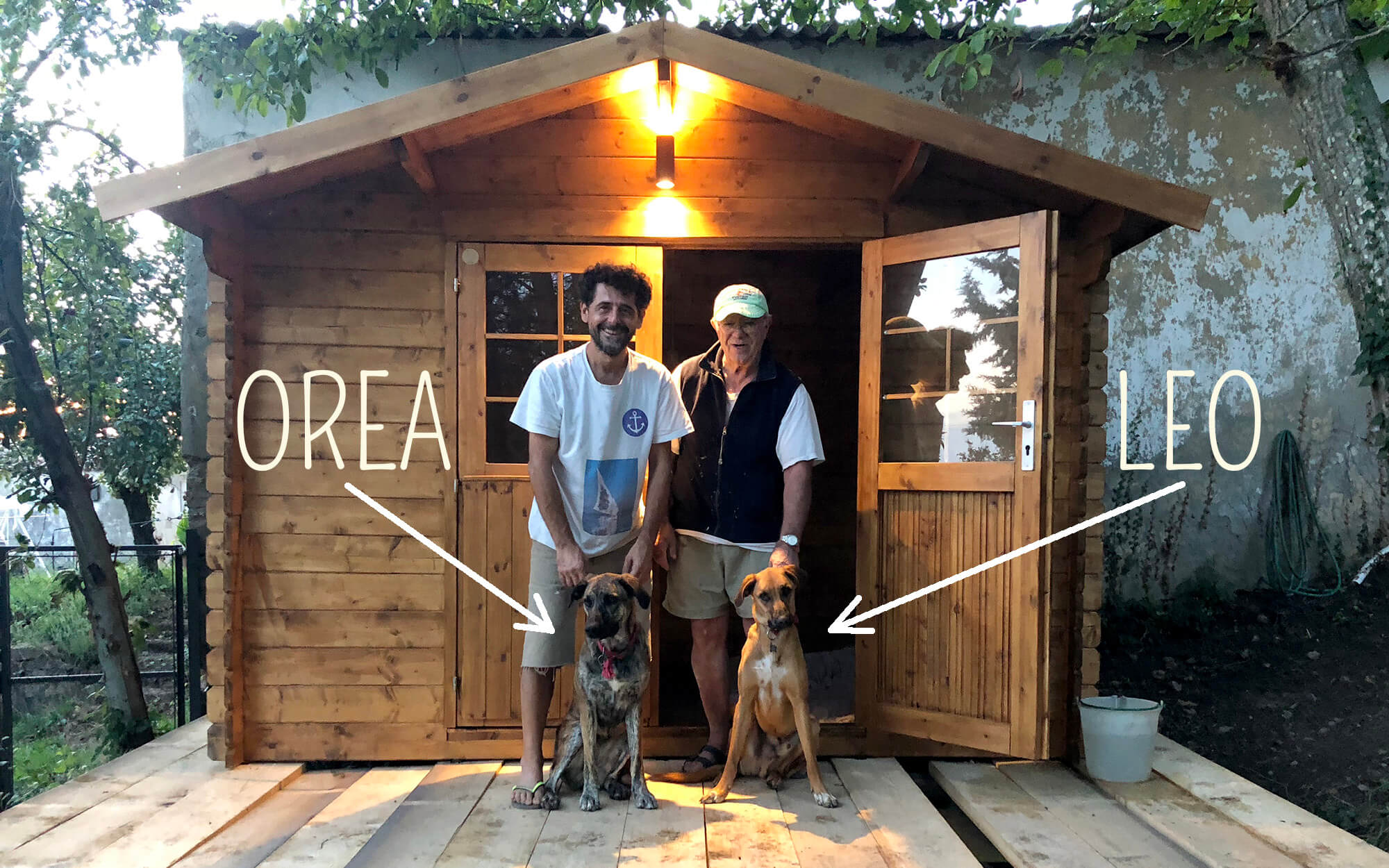 Fabrizio e Giorgio posano con i cani Leo e Orea davanti alla casetta in legno dell'area giochi per cani al Podere Camaiano